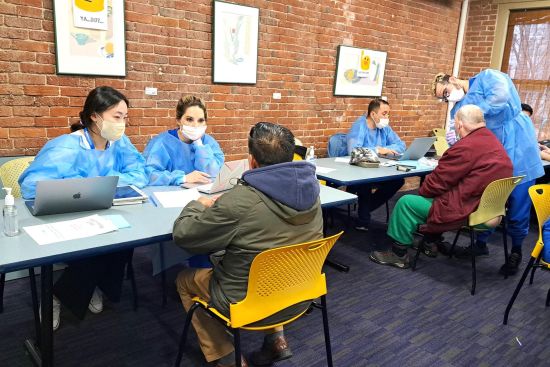 Dental students wearing gowns and masks sit across from members of the Boston Living Center; one student is examining a patient's mouth. Dental students wearing gowns and masks sit across from members of the Boston Living Center; one student is examining a patient's mouth.