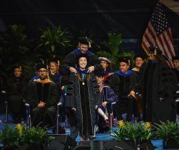 Ivy phung stands on a stage wearing her black graduation robes. People sit on chairs behind her. Shane Crotty stands behind her to put a blue hood around her neck.