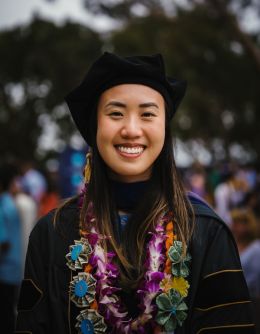 Ivy Phung faces the camera and smiles. She is wearing a black gradulation robe and cap. she is wearing a necklace of flowers and a necklace of dollar bills.
